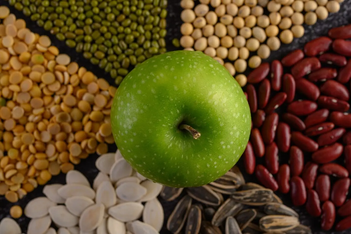 Legumes and an apple on the surface of a black cement floor.