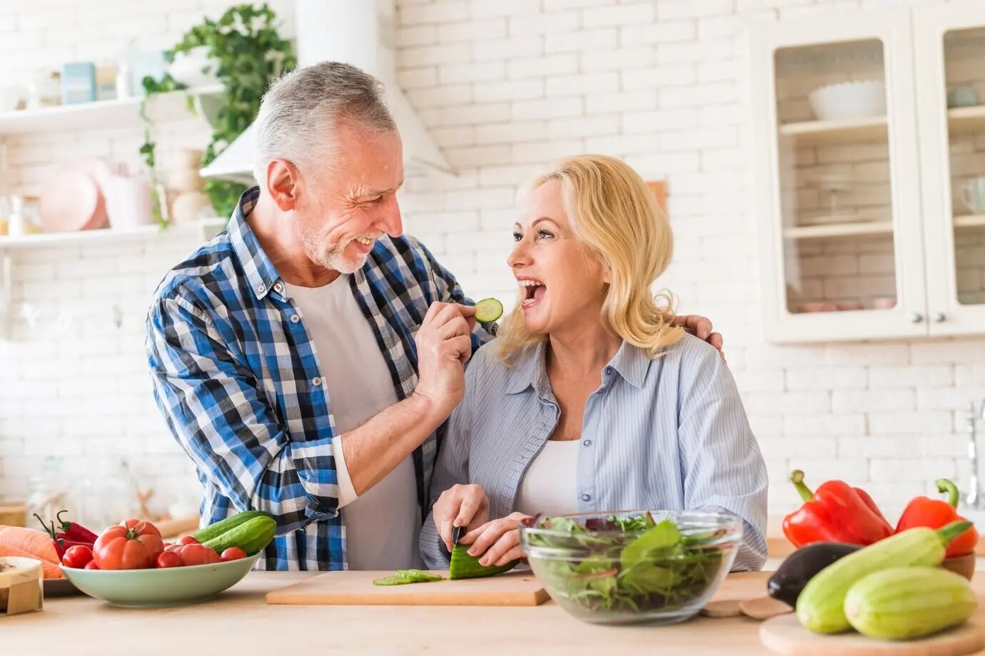 An elderly man feeds his wife a slice of cucumber in the kitchen.