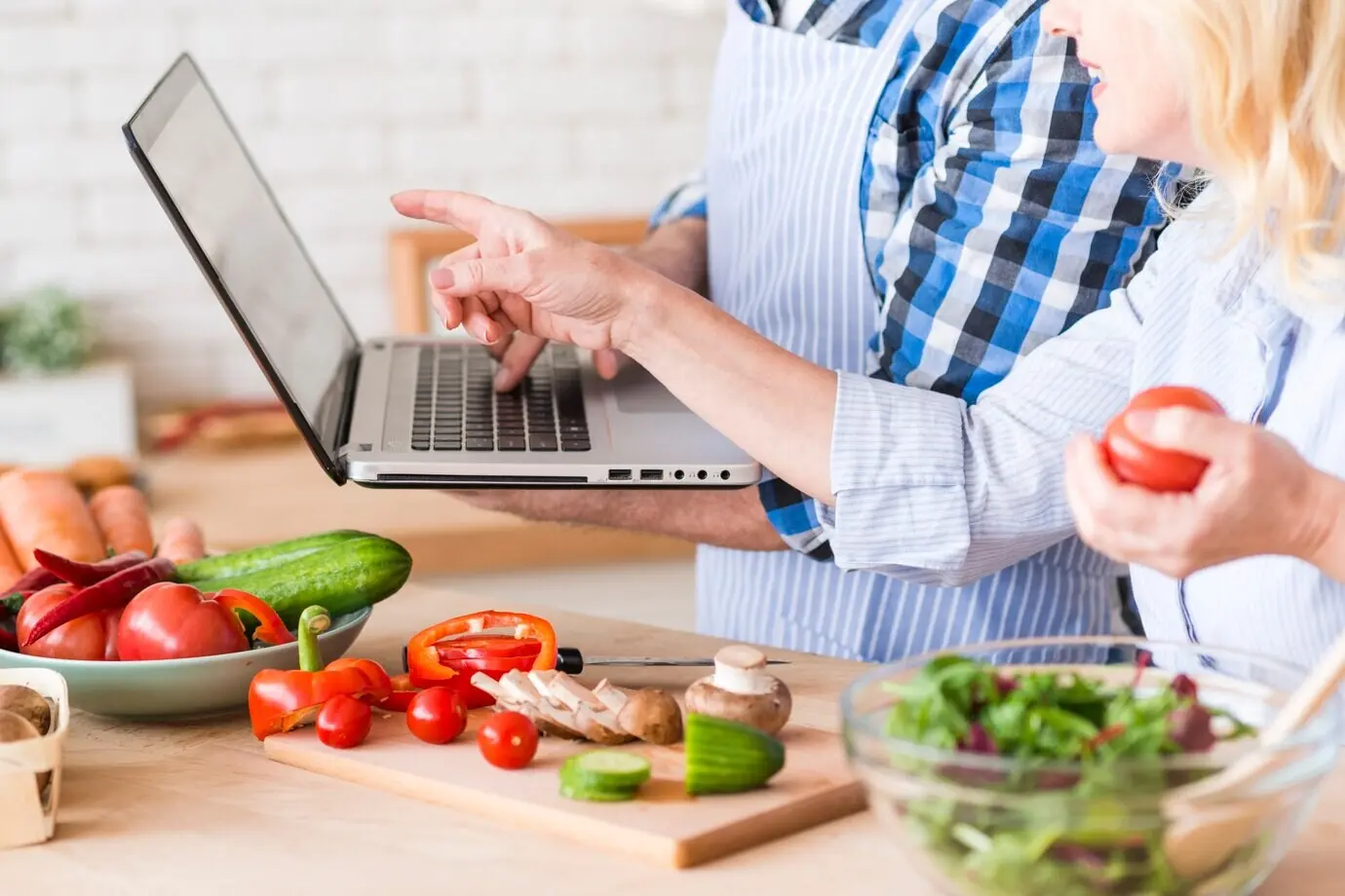 Close-up of a senior woman pointing at a laptop held by her husband while preparing the vegetable salad.